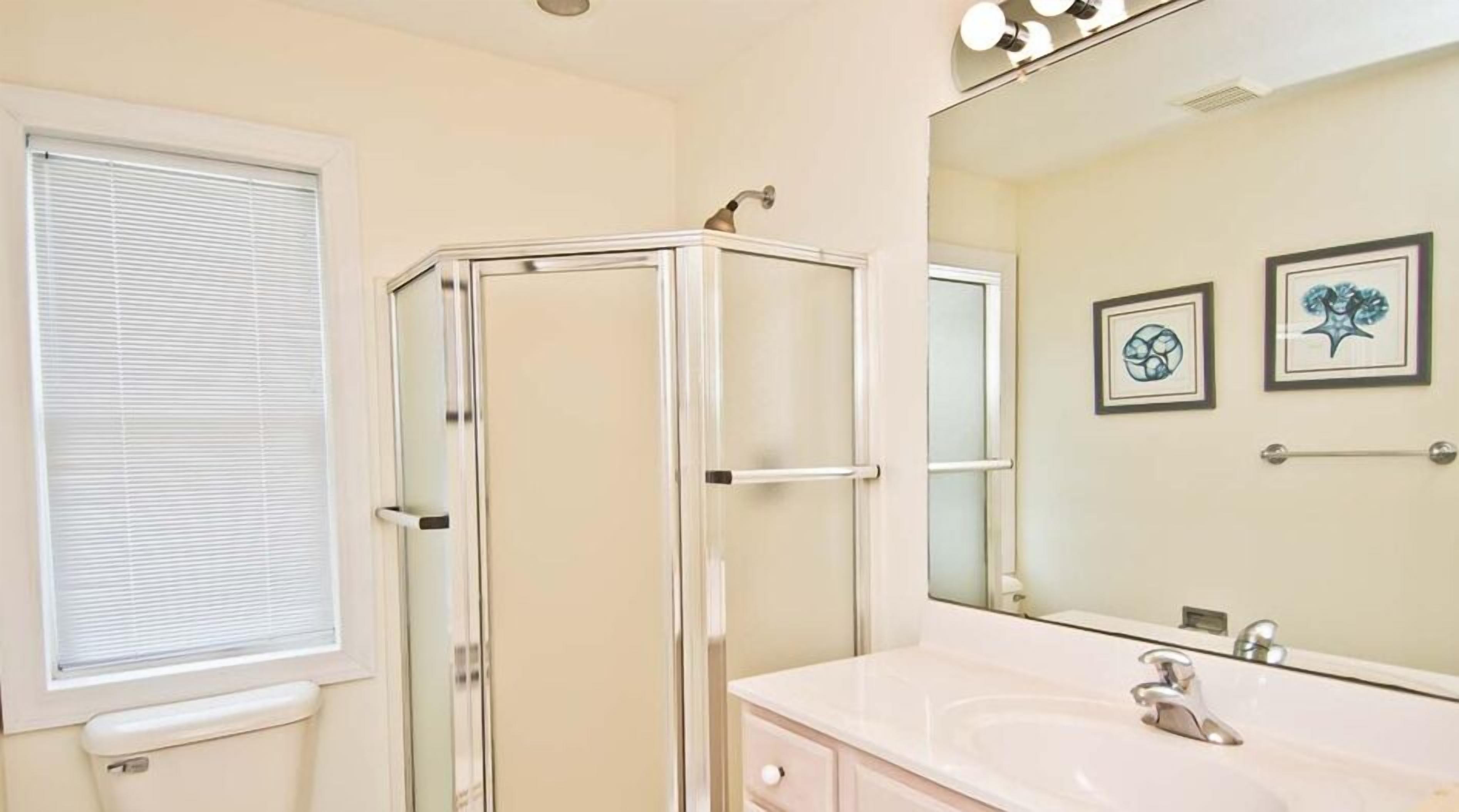 A bathroom with a glass shower enclosure, a sink with a mirror, and two framed artworks on the wall.