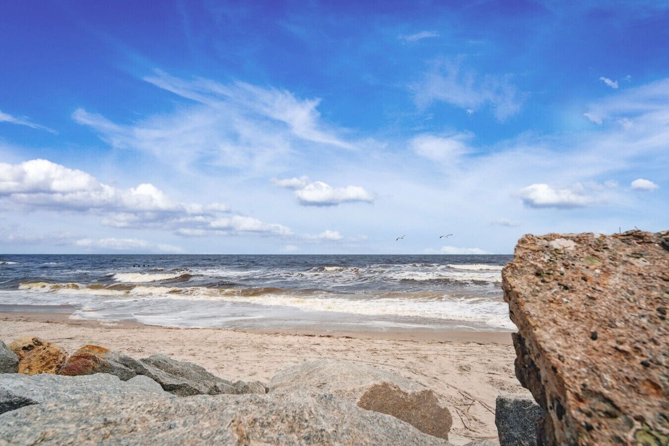 A beach with waves, a clear sky, and a large rock in the foreground.