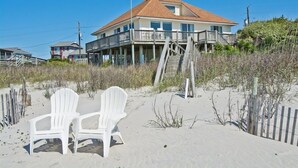 Une maison située en bord de mer, dotée d’un balcon, de deux chaises blanches sur le sable et d’une clôture en bois.