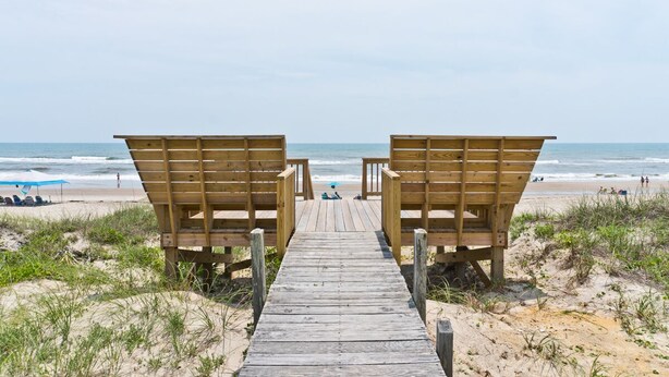 Una passerella in legno conduce a sedie a sdraio su una spiaggia sabbiosa, con l'oceano sullo sfondo.