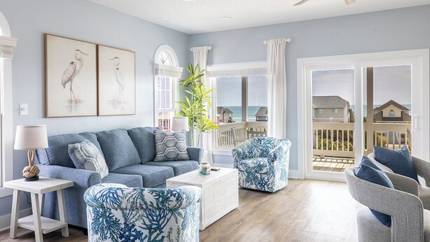 A spacious living room with a blue sofa, matching armchairs, and a coffee table. A ceiling fan and a large window with a view of the ocean are visible.