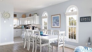 A bright kitchen with white cabinets, a dining table with chairs, and large windows.