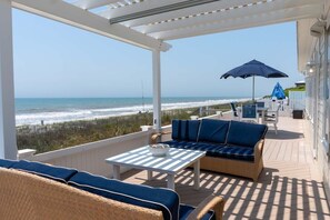 A covered outdoor seating area with a view of the beach, white furniture, and a blue umbrella.