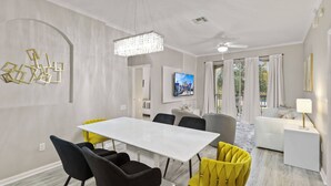 Ein modernes Wohnzimmer mit einem weißen Esstisch, schwarzen und gelben Stühlen, einem grauen Sofa und einem weißen Beistelltisch mit Lampe.