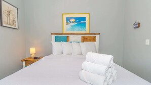 Una cama bien hecha con sábanas blancas, cabecera de madera con un panel decorativo, un cuadro enmarcado sobre la cama y una mesita de noche con una lámpara y control remoto.