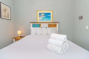 Una cama bien hecha con sábanas blancas, cabecera de madera con un panel decorativo, un cuadro enmarcado sobre la cama y una mesita de noche con una lámpara y control remoto.