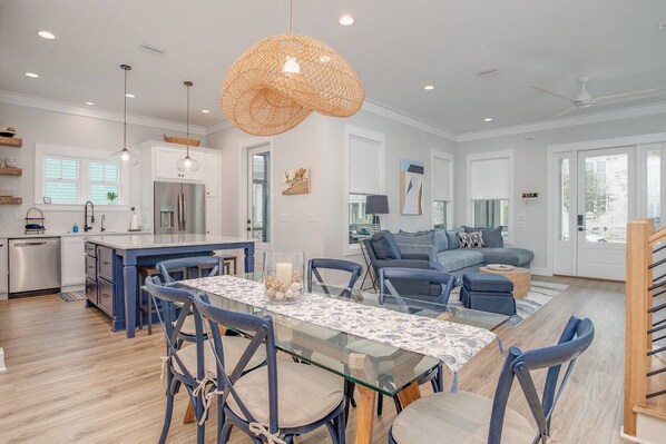 A modern kitchen with a central island, dining area with a glass table, and a living room with a sofa and a ceiling fan.