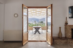 An open door leading to a patio with a table and chairs, a fireplace, and a view of a landscape with mountains. - Theodora's Family Suite (Paros)