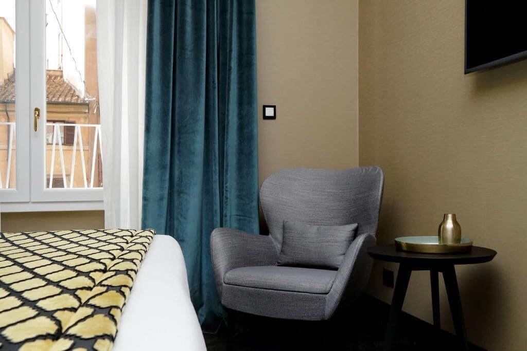 Quarto de hotel com uma cama, uma poltrona cinzenta, uma mesinha com um vaso e uma TV fixada na parede.