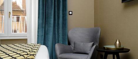 Quarto de hotel com uma cama, uma poltrona cinzenta, uma mesinha com um vaso e uma TV fixada na parede.