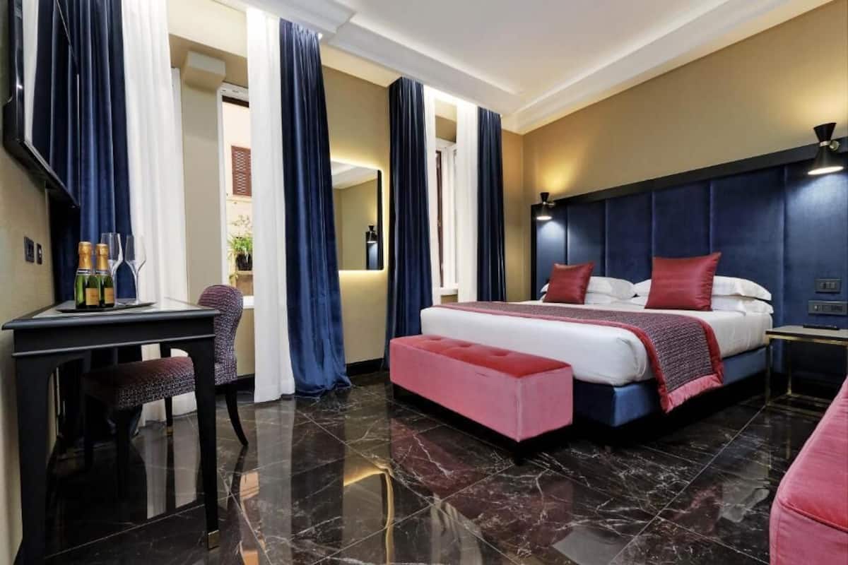 Habitación de hotel con una cama grande, un escritorio con silla, una mesita con botellas y un reposapiés rojo.