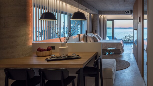 Una habitación de hotel moderna con una mesa de comedor de madera, dos lámparas colgantes negras, un ventanal con vistas al mar y una zona de estar acogedora con un sofá y una pequeña mesa redonda.
