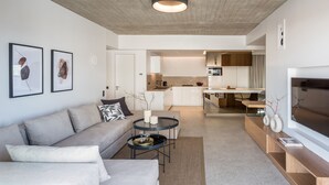 A modern living room with a sofa, coffee table, and TV. There is a kitchen in the background with cabinets and a dining area.