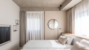 A hotel room with a large bed, a round mirror, a TV, and a window with curtains.
