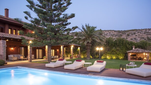 Ein Poolbereich mit Liegestühlen und einem kleinen Pool, umgeben von einem angelegten Garten und einem Gebäude mit Außenbestuhlung.