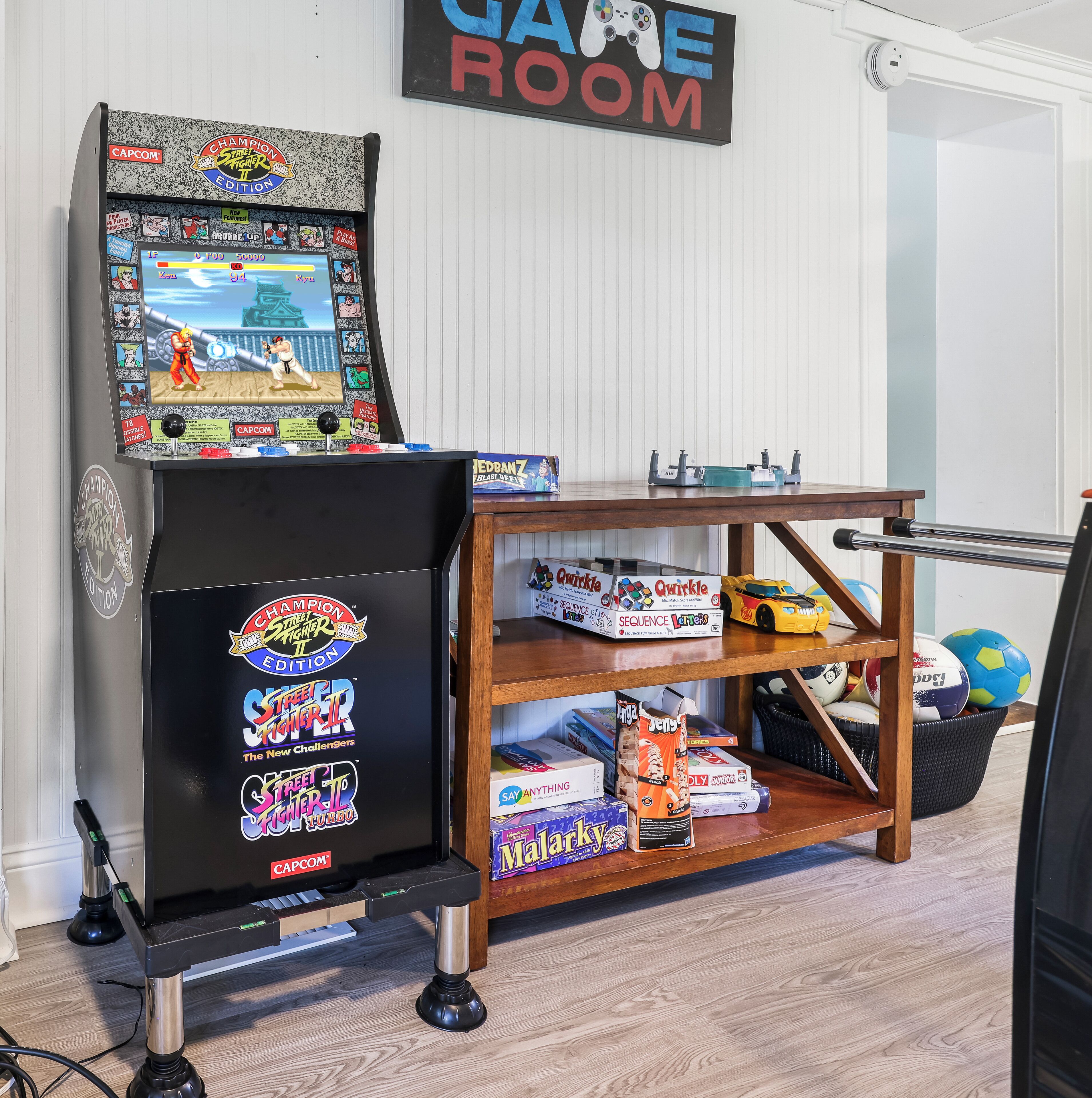 En spillearkade med et skilt der siger 'Game Room', en arkademaskine 'Champion Street Fighter II Edition' og en reol med forskellige legetøj og spil.