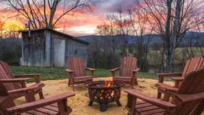 Un patio in legno con quattro poltrone Adirondack attorno a un braciere centrale, sullo sfondo un deposito di legno e un tramonto su un paesaggio collinare.