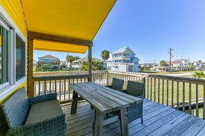 A wooden deck with a table and chairs, a yellow awning, and a view of houses in the background. - Hello Sunshine by Ryson Vacation Rentals (Galveston)