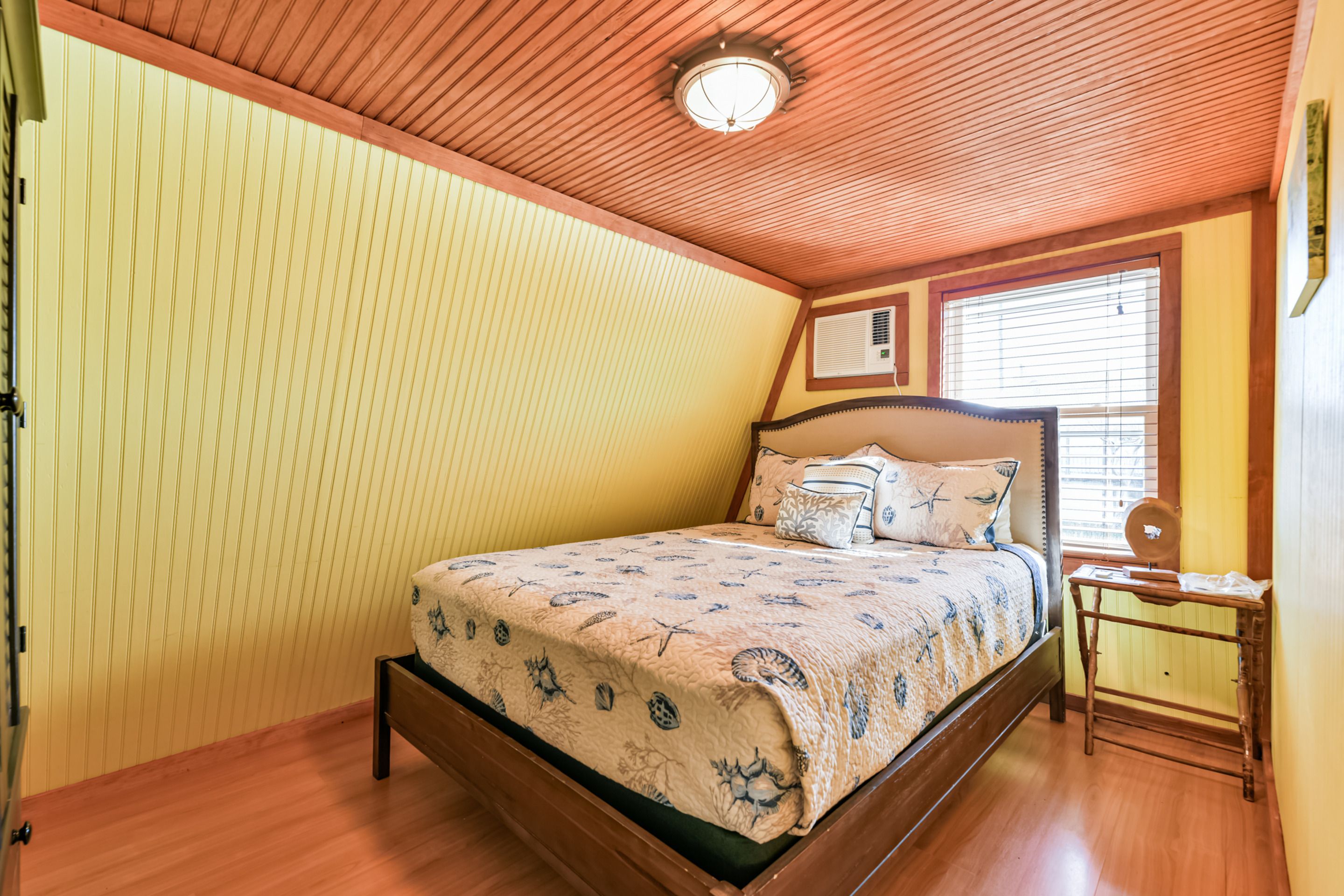 A bedroom with a wooden ceiling, a bed with a patterned bedspread, a bedside table, and a window with blinds.