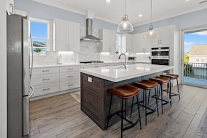 A modern kitchen with white cabinetry, a large island with stools, a stainless steel range hood, and pendant lighting.