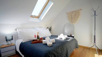 Een net opgemaakt bed met een donkere deken, een nachtkastje met een lamp, een dienblad met wijn en glazen, en een hoge vloerlamp.
