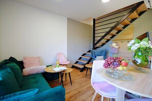A modern living room with a green sofa, a small round table with a book and a bottle, a staircase with wooden steps, and a dining area with a glass table set with fruit.