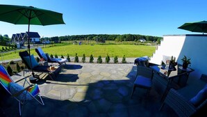 A patio with lounge chairs, a table, and umbrellas, overlooking a grassy field and a house.