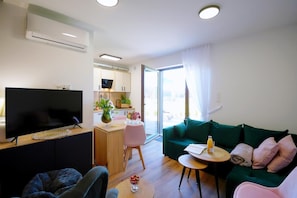 A modern living room with a flat-screen TV, a green sofa, a dining area with a pink chair, and a kitchen in the background.