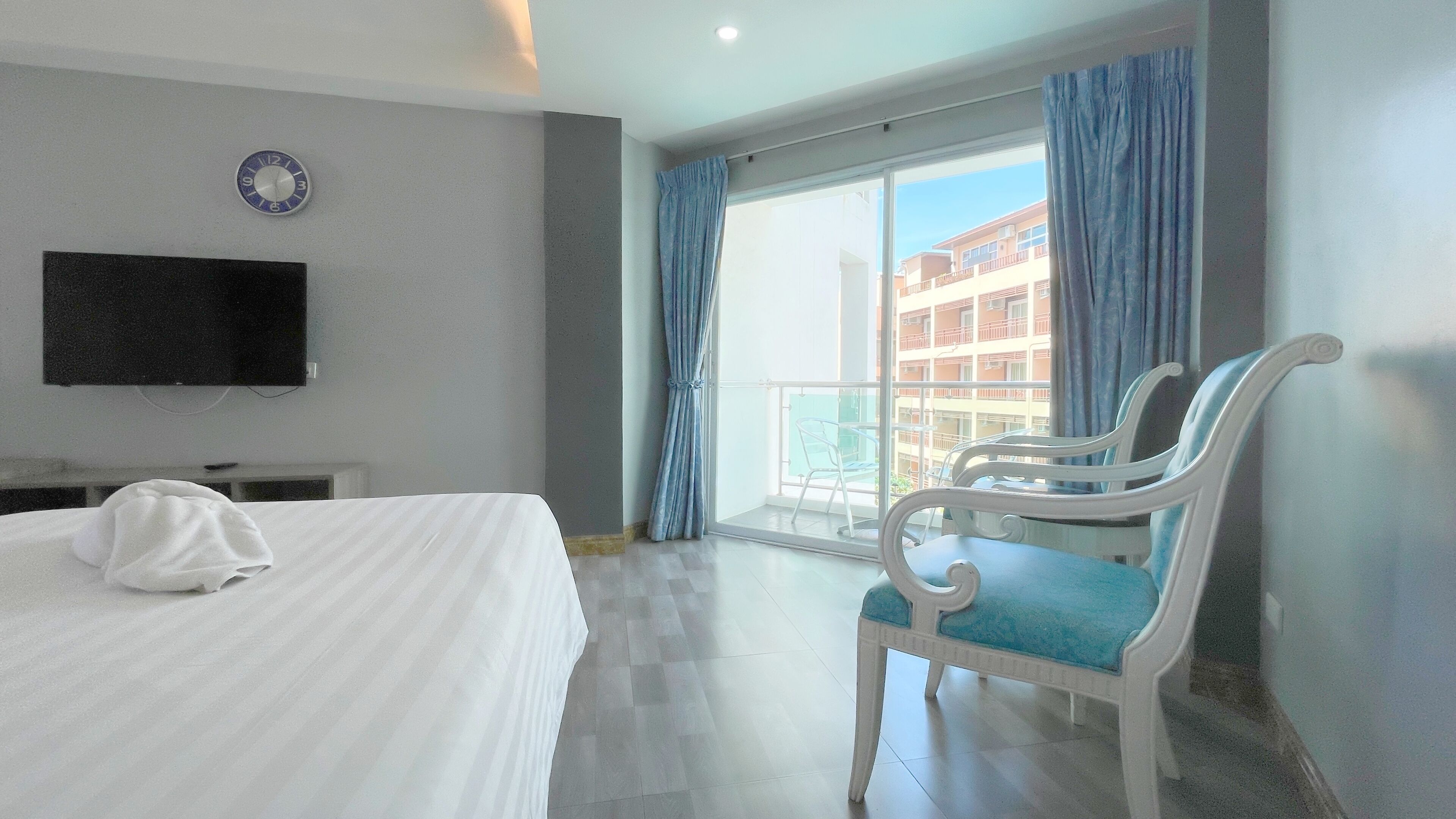 A hotel room with a bed, a TV mounted on the wall, a balcony with blue curtains, and a chair with a blue cushion.