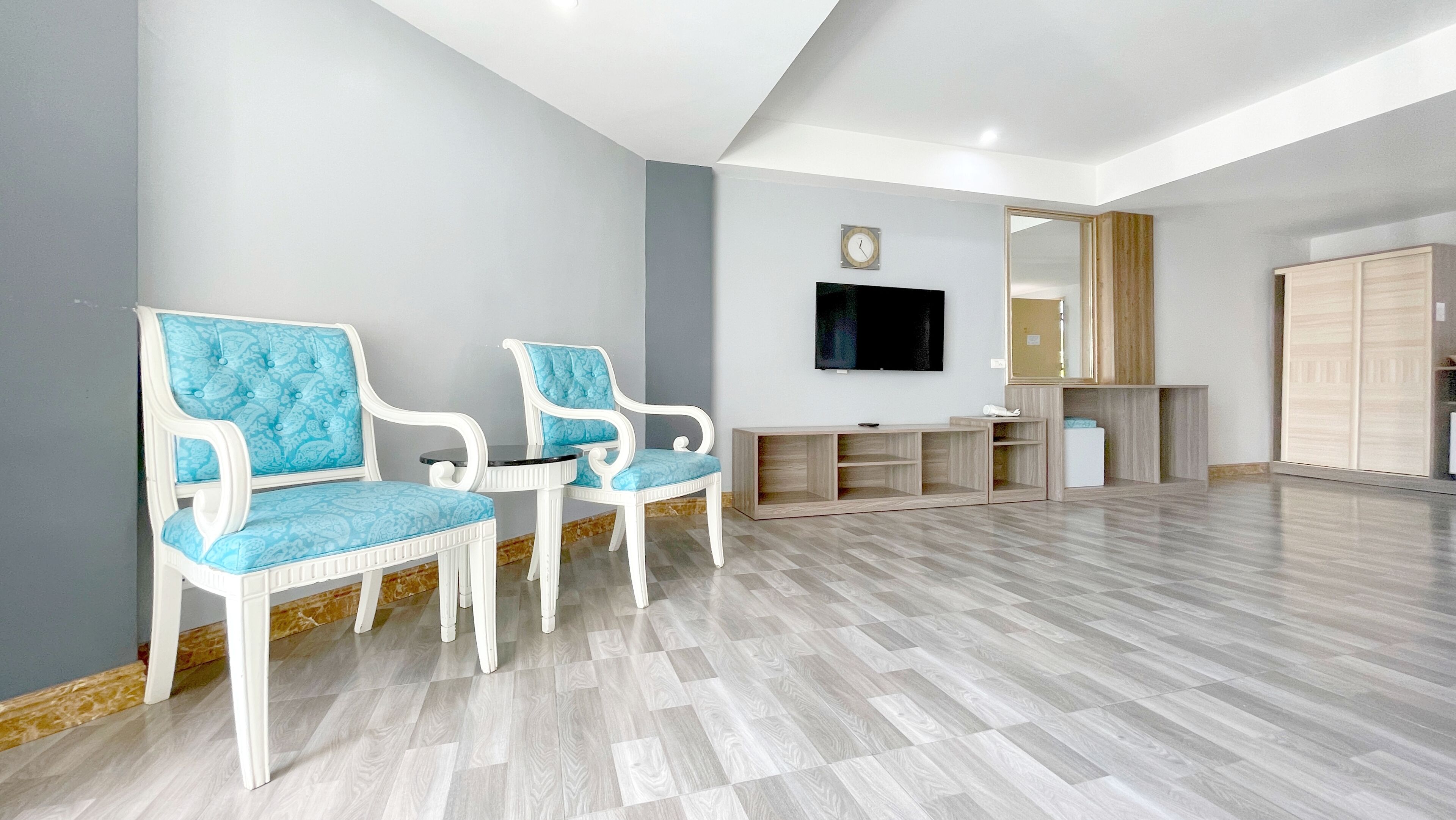 A spacious room with a flat-screen TV mounted on the wall, two white chairs with blue cushions, a wooden cabinet, and a wardrobe.