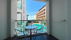 Un balcón con barandilla de vidrio, una mesa pequeña y dos sillas, con vista a una piscina y un edificio con varios balcones.
