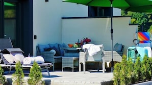Un espace extérieur moderne, aménagé avec des fauteuils de détente, un canapé et une table surmontée d’un parasol vert.