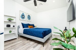 Un dormitorio moderno con una cama, paredes blancas, un ventilador de techo y un televisor montado en la pared.