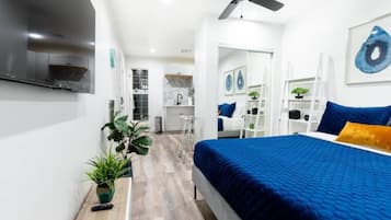 Une chambre moderne avec un grand lit, un ventilateur de plafond, un téléviseur fixé au mur et, au fond, une petite cuisine.