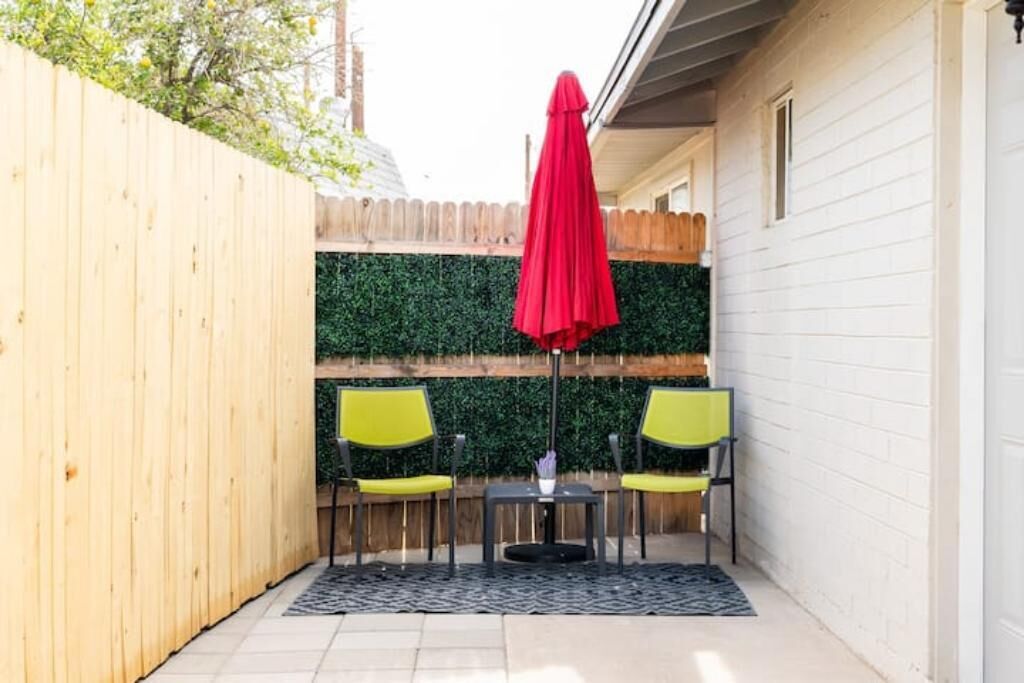 A small patio with a wooden fence, a green hedge, and two yellow chairs with a red umbrella.