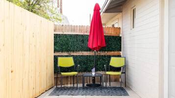 A small patio with a wooden fence, a green hedge, and two yellow chairs with a red umbrella.