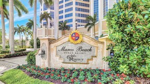 A sign for Marco Beach Ocean Resort with a crest emblem, surrounded by landscaped greenery and palm trees. - Marco Beach Ocean Resort Unit 712 (Marco Island)