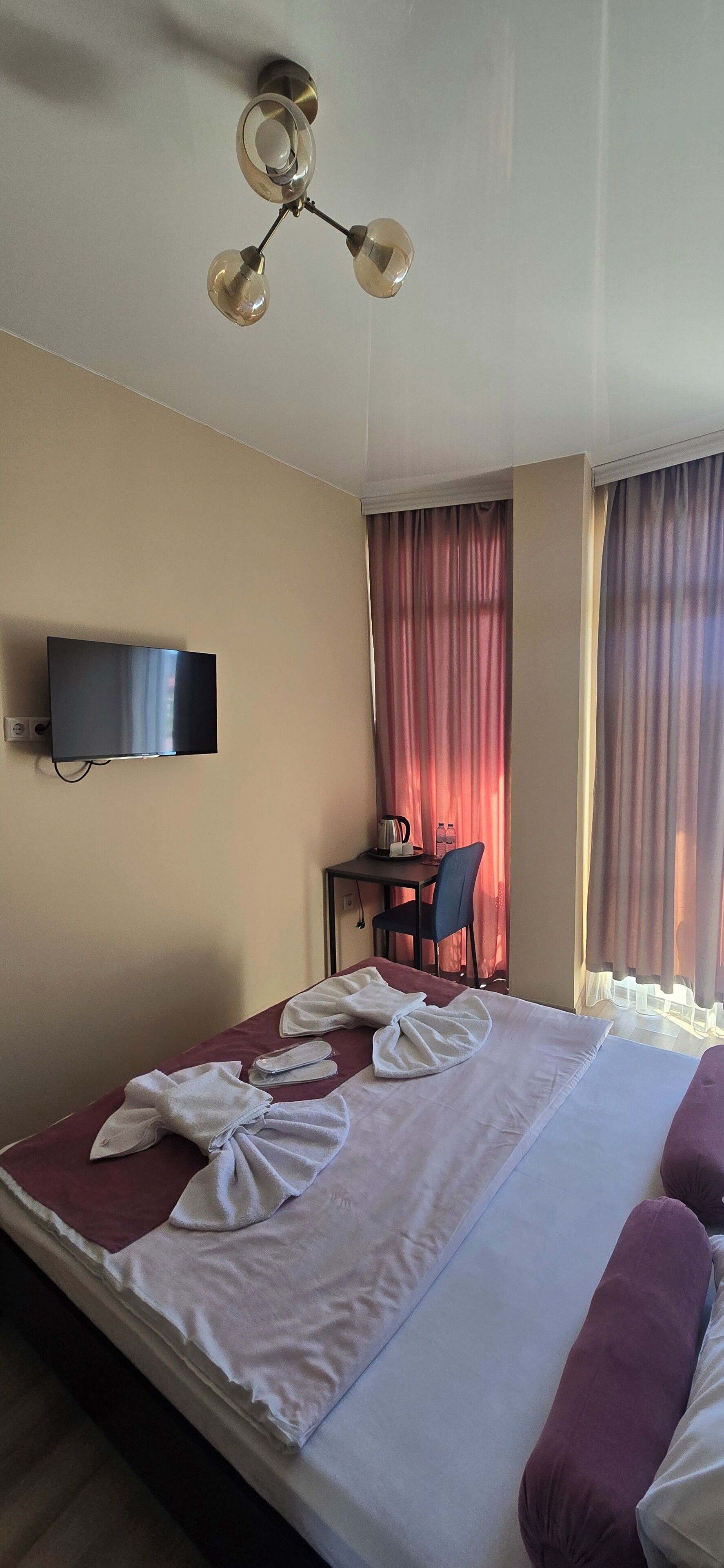 Ein Hotelzimmer mit einem Bett, einem Schreibtisch, einem Stuhl, einem Fernseher und Vorhängen.