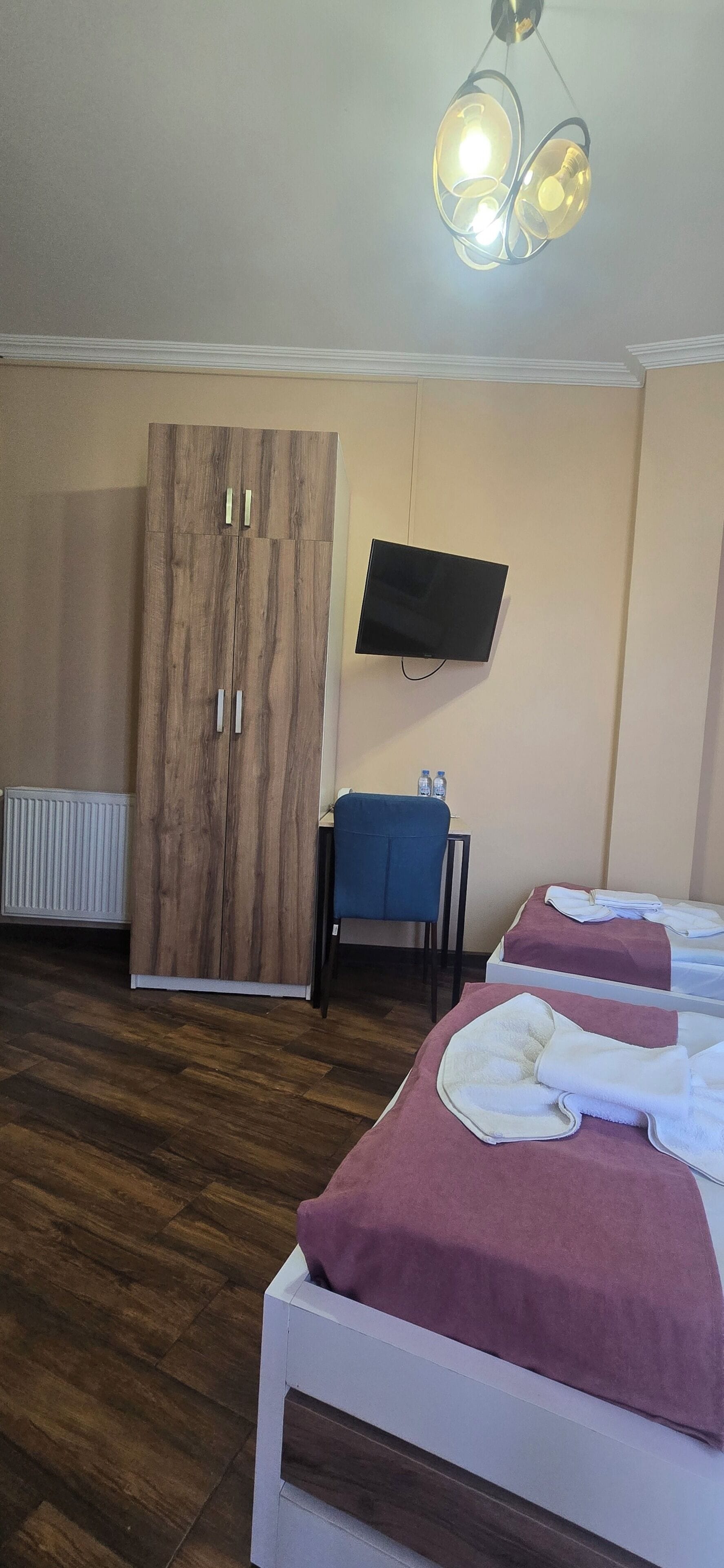 A hotel room with a wooden wardrobe, a television mounted on the wall, two beds with towels, a chair, and a wooden floor.
