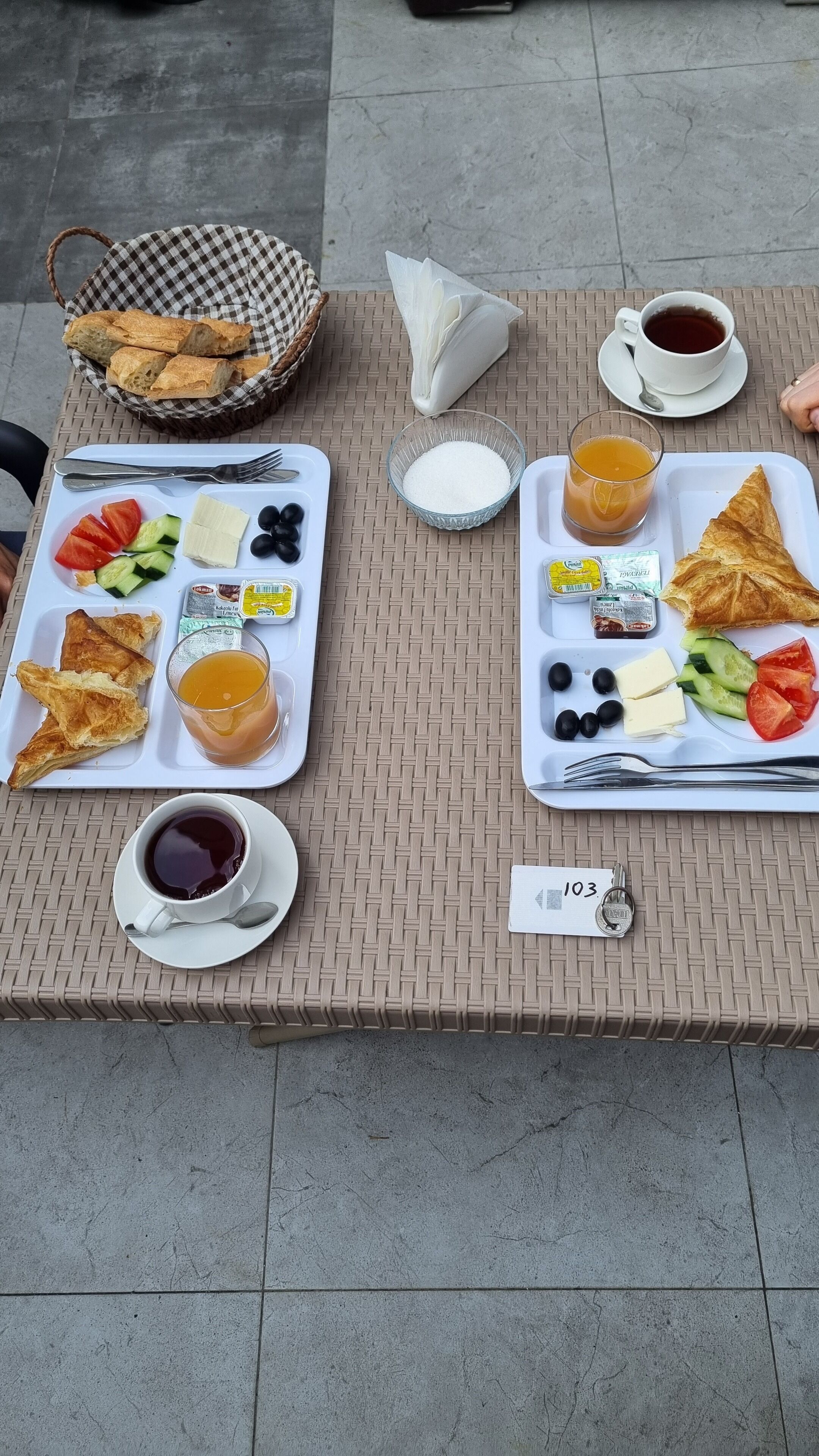 A breakfast setup with two trays of food, including bread, cheese, tomatoes, cucumbers, olives, and beverages.
