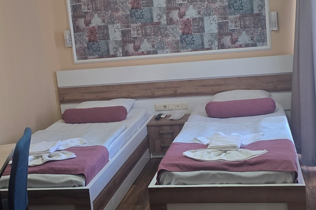 A hotel room with two beds, a wooden floor, a ceiling light fixture, and a large patchwork wall hanging.