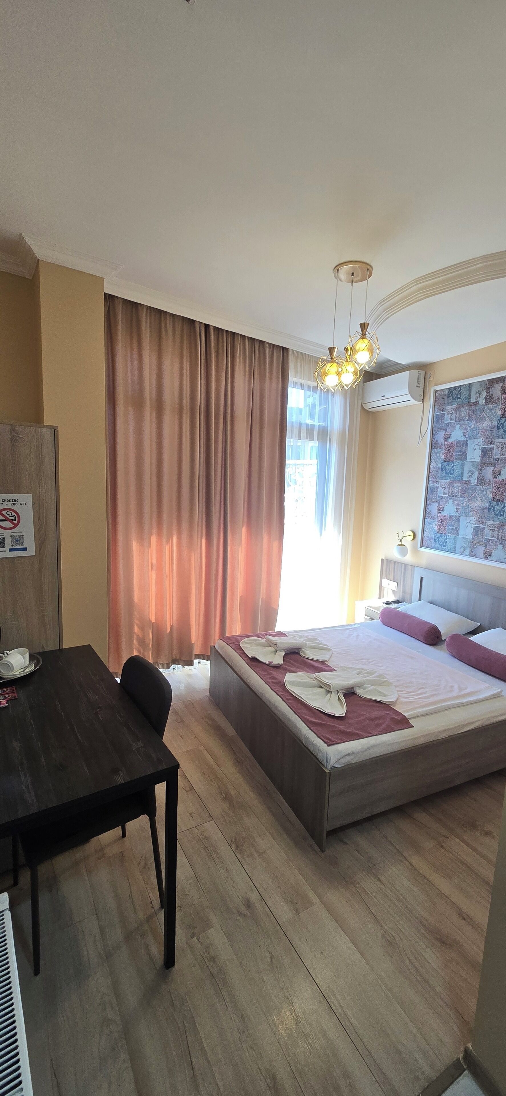 A hotel room with a wooden floor, a bed with a red and white bedspread, a desk with a chair, a dining table, and a large window with curtains.