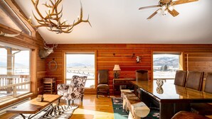 Ein Holzblockhaus-Interieur mit einem großen Holzesstisch, Ledersesseln, einem Zebra-Teppich und einem Hirschkopf an der Wand.