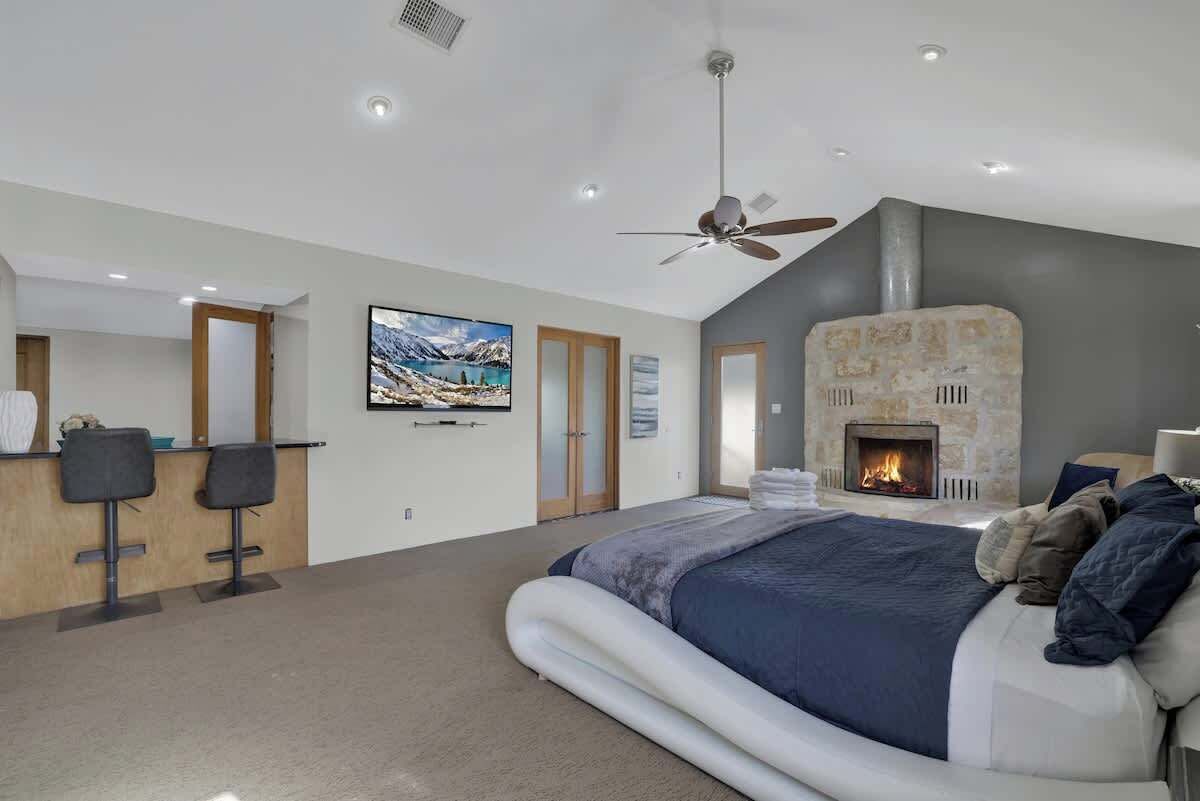 A bedroom with a bed, a fireplace, a ceiling fan, and a bar area with two stools.