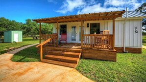 Une terrasse en bois mène à un porche couvert, accessible par un escalier. On y trouve également un petit abri de jardin et une maison avec un toit en métal.