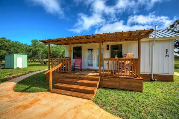 A wooden deck leading to a covered porch with a staircase, a small shed, and a building with 'ELM' on the side.