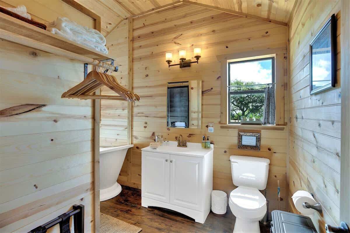A wooden cabin bathroom with a white sink, toilet, and hanging clothes rack.