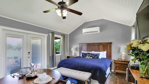 Une chambre avec un grand lit, un ventilateur de plafond, une table à manger avec des chaises et une fenêtre avec des stores.