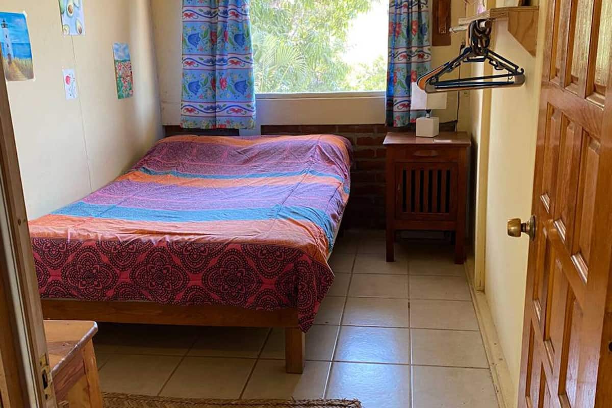 A small bedroom with a bed, a wooden desk, a hanging clothes rack, and a door.