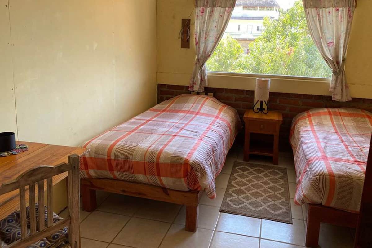 A small, cozy room with two single beds, a wooden bench, a small table, and a window with curtains.